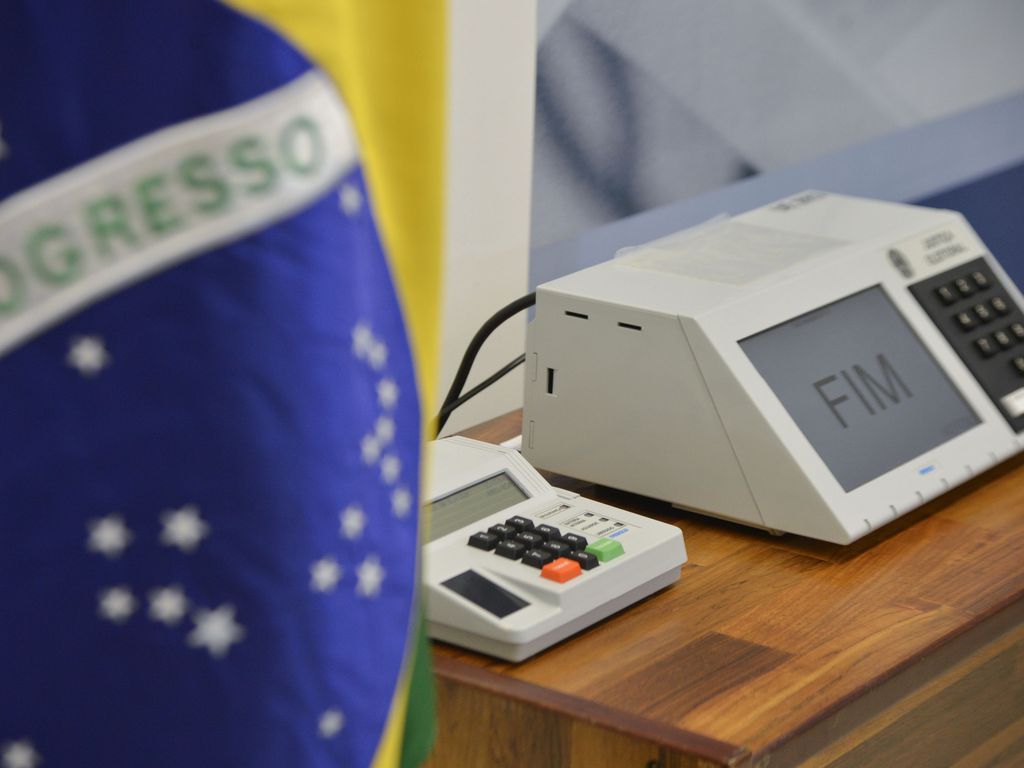 Urnas eletrônicas ao lado de bandeira do Brasil em seção eleitoral do TSE