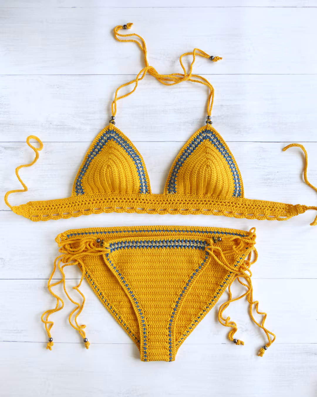 Handmade yellow crochet bikini set laid flat on white wooden surface — mustard cotton two-piece with scalloped edges and tie straps
