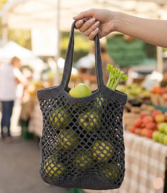 Hand holding a grey crocheted mesh market bag filled with green apples and celery at a farmers market