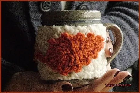 Crochet cup cozy with orange maple leaf appliqué on ceramic mug, autumn cottagecore craft