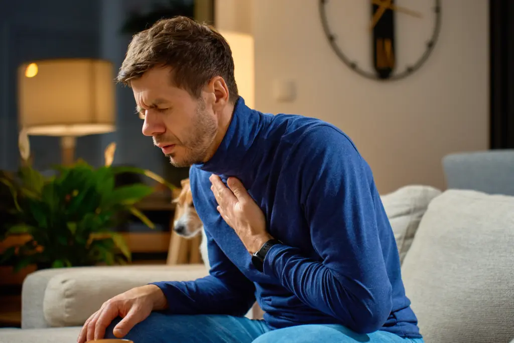 Man sitting on sofa holding his chest and throat experiencing acid regurgitation and heartburn