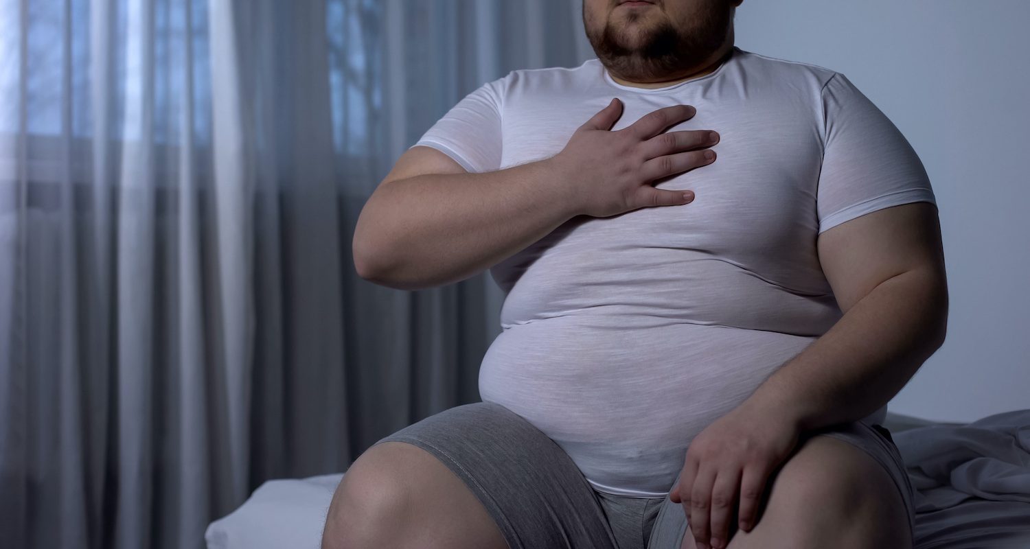 Overweight man sitting on bed holding his chest showing how excess abdominal weight causes acid reflux