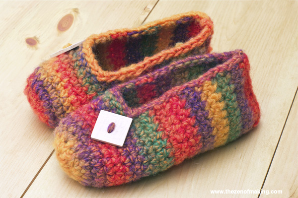 Two handmade crochet slipper styles on wooden table — rainbow granny square slippers and textured blue-gray cable knit slippers with yarn balls