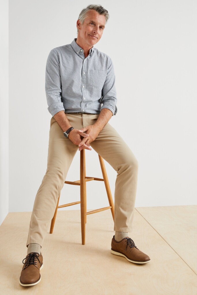 Mature man in blue checkered dress shirt with khaki chinos sitting on stool showing relaxed professional style